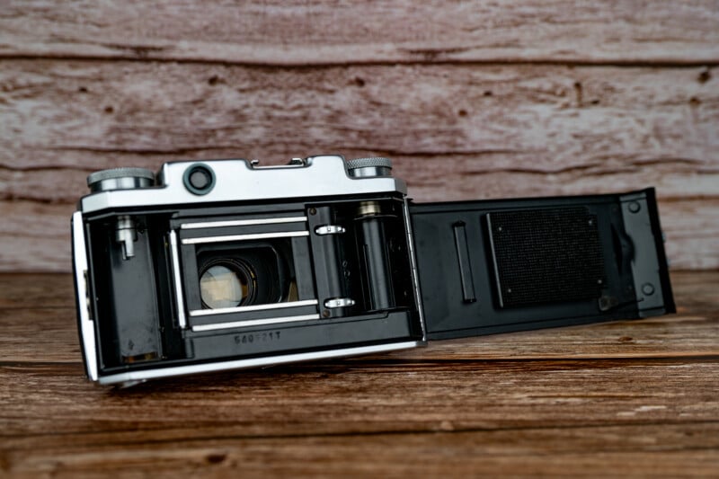 A vintage 35mm film camera with its back door open, revealing the internal film chamber and shutter mechanism, resting on a wooden surface.