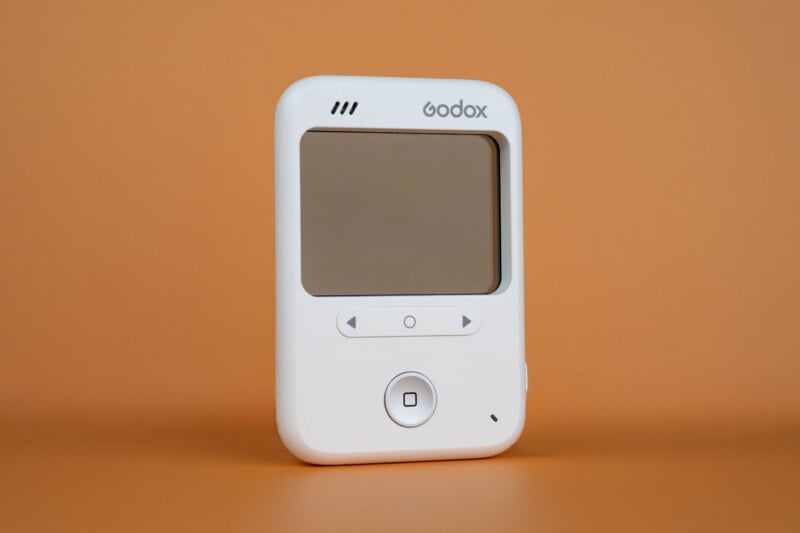 A white Godox wireless flash trigger with a blank screen, directional buttons, and a central circular button, set against a solid orange background.