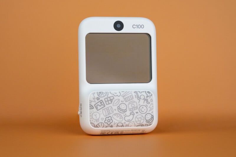 A white, rectangular Godox C100 transparent viewfinder camera with a patterned lower section sits centered against a solid orange background.