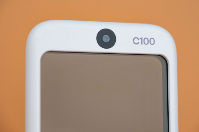 A close-up of the top portion of a white electronic device featuring a centered circular camera lens and the text "C100" printed to its right against a solid orange background.