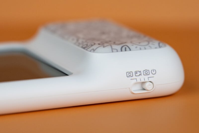 A close-up of a white device with a sliding switch, showing icons for power, camera, video, and a square frame on an orange background.