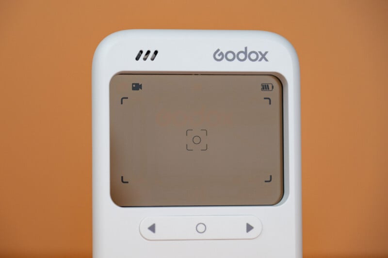 A close-up of a white Godox device with a screen displaying a video icon, a battery indicator, and a central focus frame, positioned against a solid orange background.