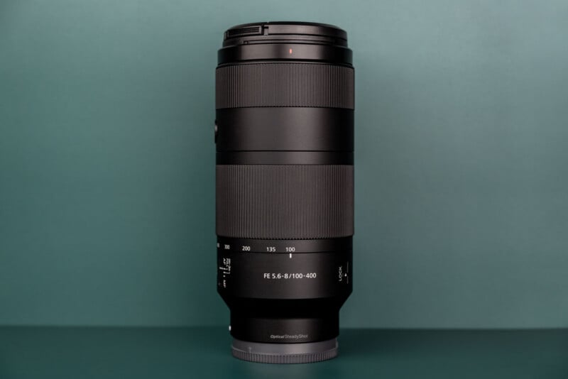 A black telephoto zoom lens stands vertically against a solid teal background, with text on the barrel reading "FE 5.6-8/100-400".