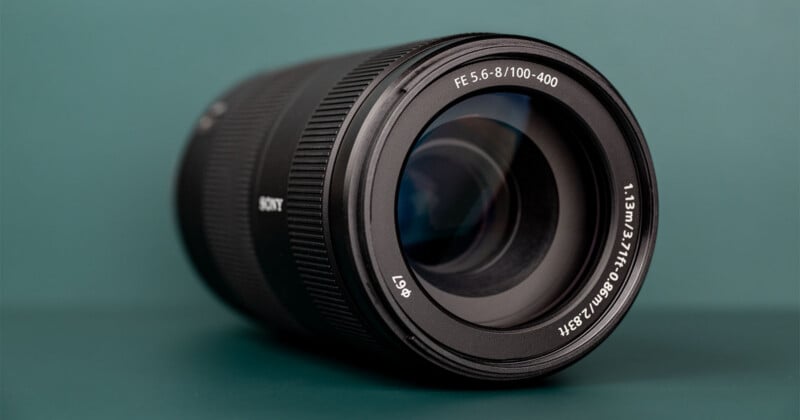 A black Sony telephoto zoom lens sits on a dark green surface, showing the front element with text reading "FE 5.6-8/100-400" and "φ67".