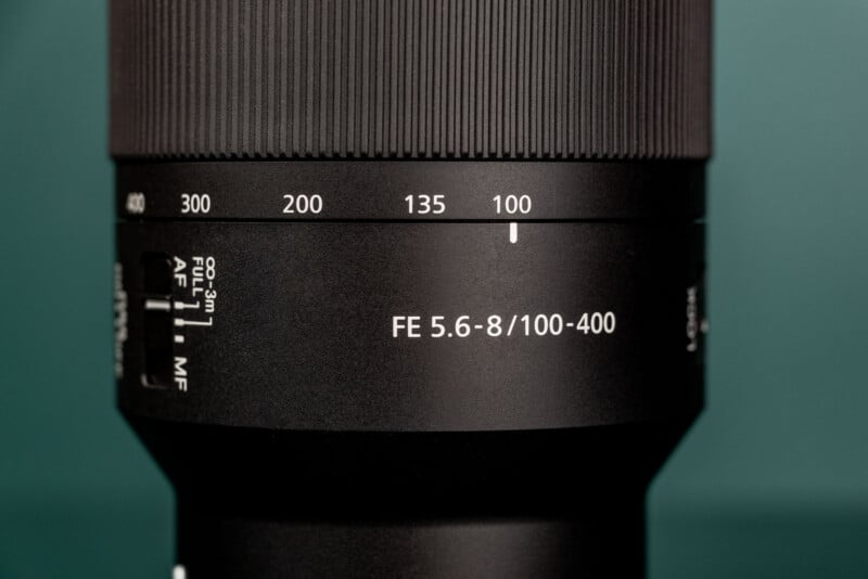 A close-up of a camera lens barrel showing a zoom ring set to 100mm, with focus and aperture markings visible.