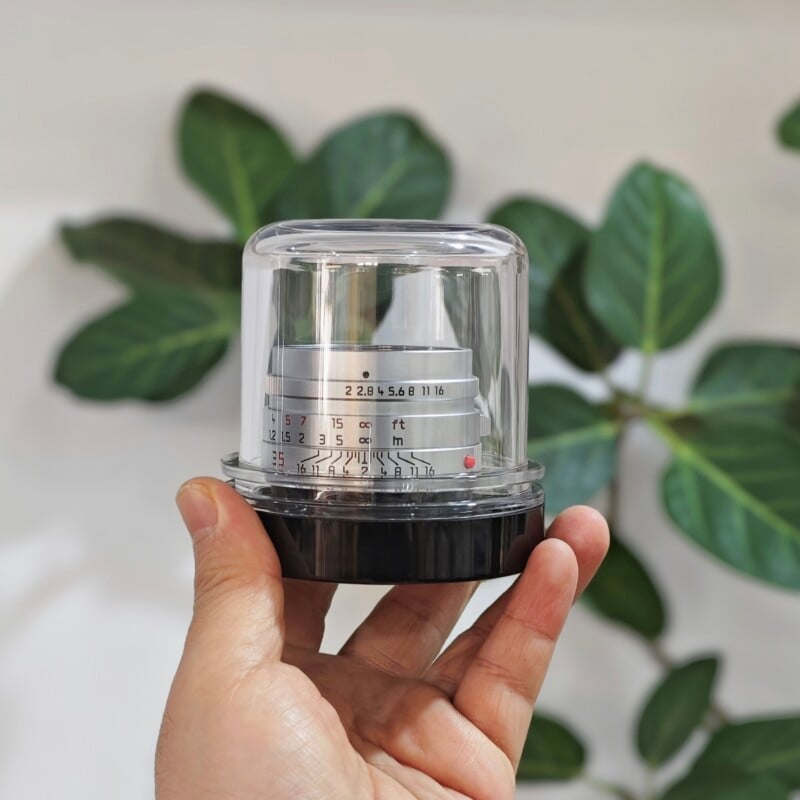 A hand holds a silver camera lens encased in a clear plastic display dome against a blurred background of green leaves.
