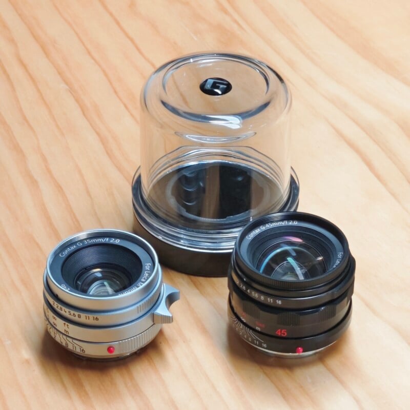 Two camera lenses and a clear plastic storage case sit on a light-colored wooden surface.