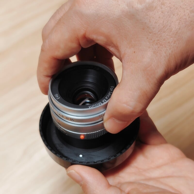 A person's hands hold a silver camera lens with a black lens cap attached, showing the text "For Leica M Mount" and "E46" on the front rim.