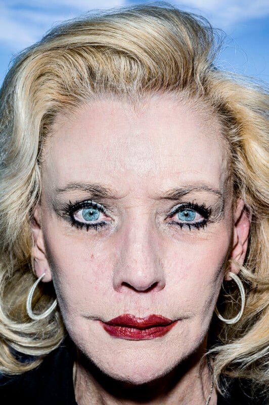 A close-up portrait of a blonde woman with blue eyes, heavy dark eyeliner, and deep red lipstick, wearing silver hoop earrings.