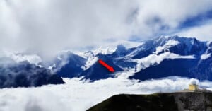 A red arrow points toward a snow-capped mountain peak rising above a thick layer of clouds, with a small building visible on a ridge in the foreground.