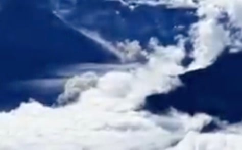 A view from above looking down at white, fluffy clouds drifting over dark, mountainous terrain.