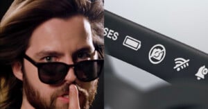 A man with long hair and a beard holds a finger to his lips while wearing sunglasses, shown next to a close-up of a device arm displaying battery, camera, Wi-Fi, and notification icons.