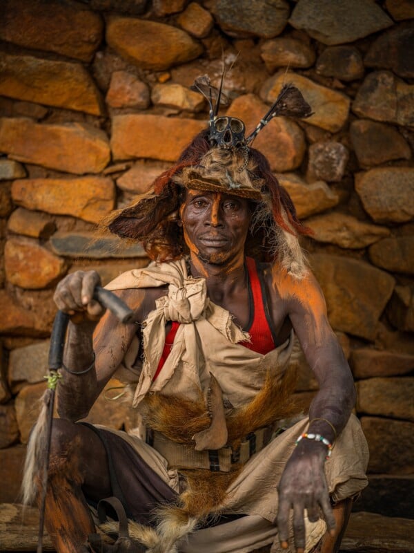 A man with orange face paint and a skull-adorned hat sits against a stone wall, wearing a red tank top and draped fabric.