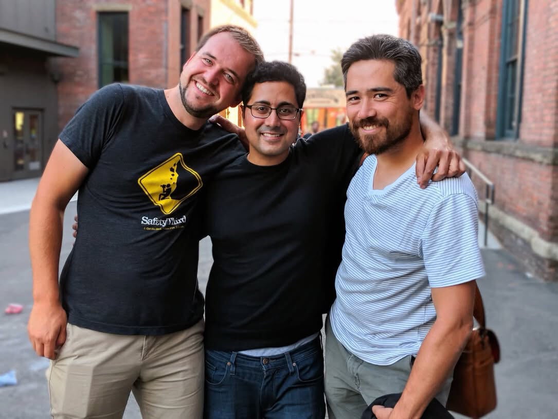 Three men stand close together outdoors with their arms around each other, smiling at the camera.