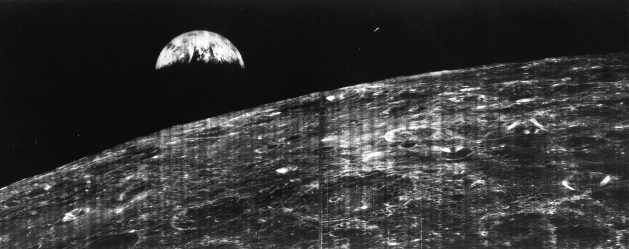 A black-and-white view of the Moon's cratered surface in the foreground with the Earth rising in the dark sky above.