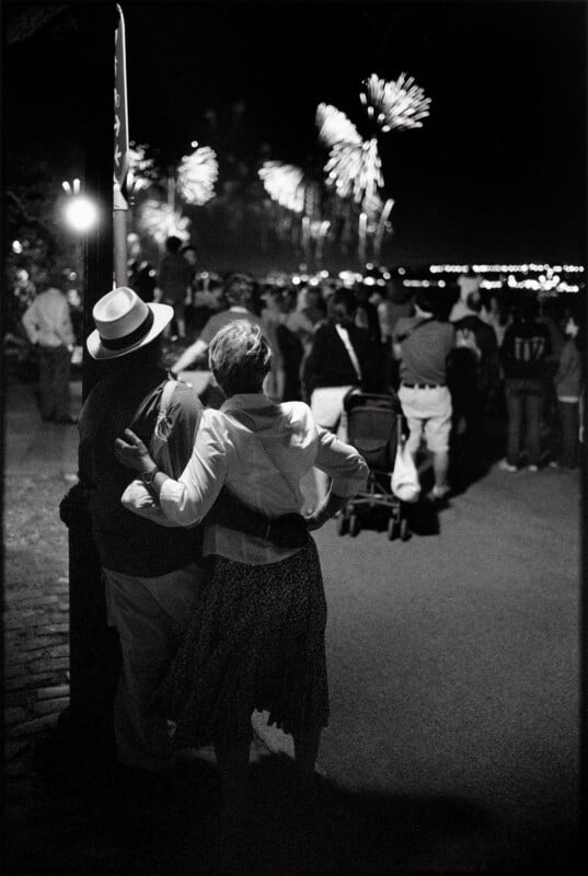 A couple stands with their arms around each other, watching fireworks over a crowd at night.