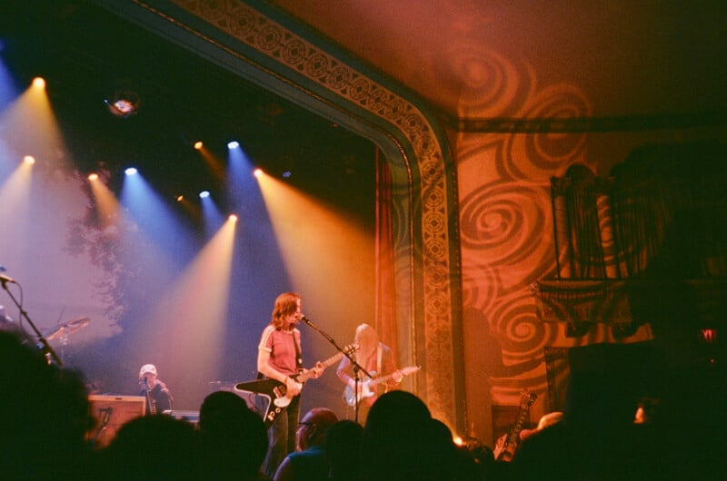 A band performs on a stage with ornate, swirling wall patterns, illuminated by blue and orange spotlights.
