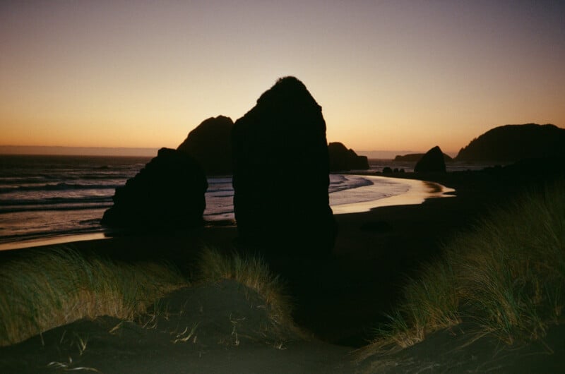 Silhouetted rock formations stand on a sandy beach at sunset, with tall dune grasses in the foreground.