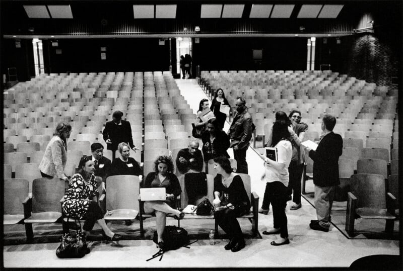 A group of people gather in the front rows of an empty auditorium, with some seated and others standing while talking.