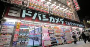 The storefront of a Yodobashi Camera store in Shinjuku at night, featuring bright neon signage, floor information displays, and rows of capsule toy machines.