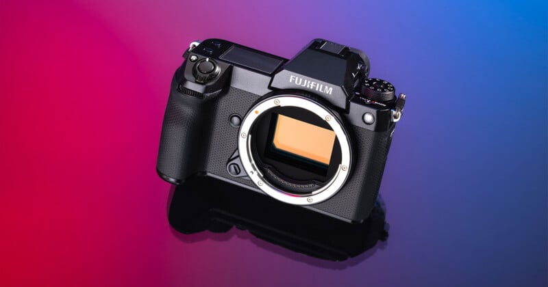 A black Fujifilm mirrorless camera body sits on a reflective surface against a gradient background transitioning from magenta to blue.