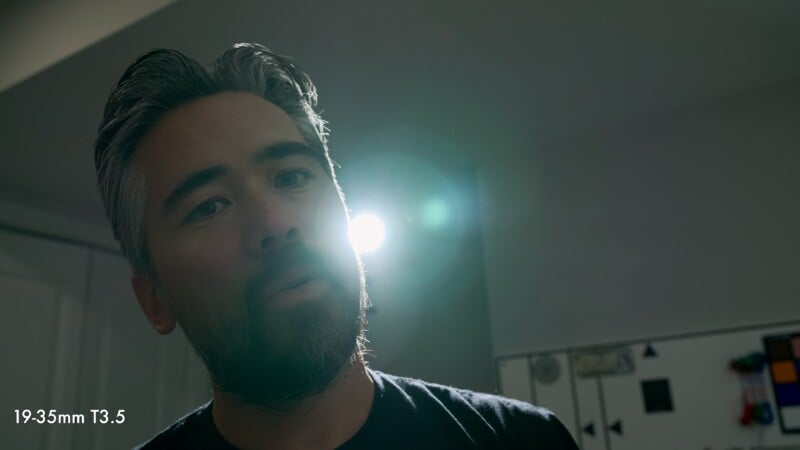 A man with graying hair and a beard looks directly into the camera, with a bright light source behind him creating a lens flare.