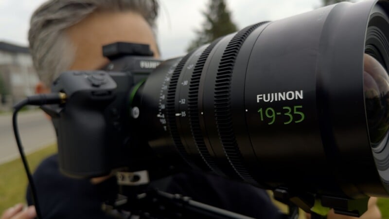 A person holds a professional camera with a large Fujinon 19-35 lens attached, focused on the lens barrel.