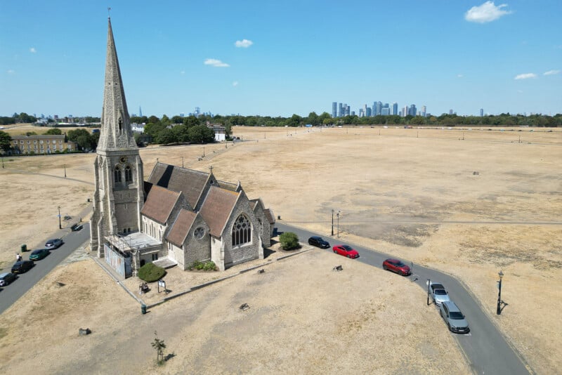 An aerial view of a stone church with a tall spire situated on a large, dry, grassy field with a line of parked cars along the road.