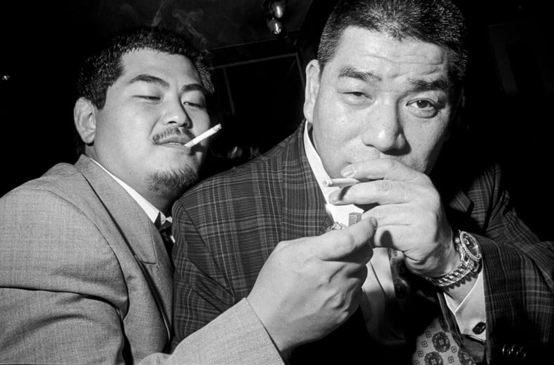 Two men in suits sit close together in a dark setting, both smoking cigarettes while one lights the other's.