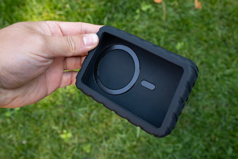 A hand holds a black, rectangular silicone protective case with a circular MagSafe-style magnetic ring and a small rectangular cutout on the interior surface.