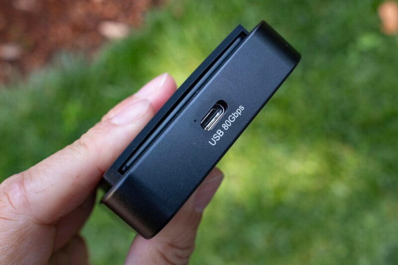 A hand holds a black electronic device featuring a USB-C port labeled "USB 80Gbps" against a blurred green grass background.