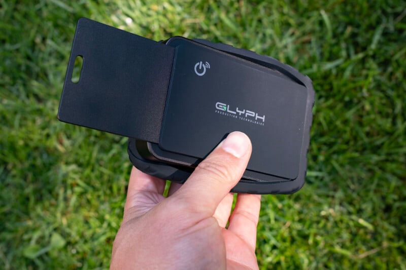 A hand holds a black Glyph Production Technologies portable hard drive with a protective rubber bumper and a partially extended black plastic card against a background of green grass.