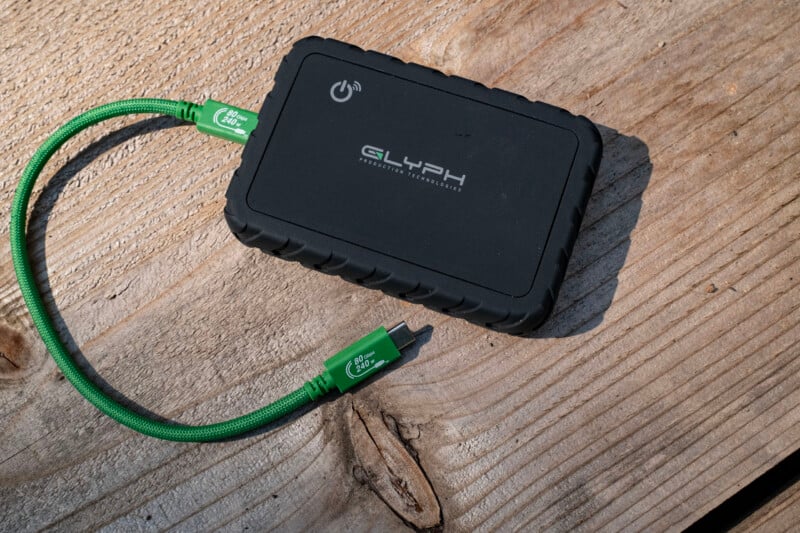 A black Glyph external hard drive sits on a weathered wooden surface, connected to a bright green USB-C cable.
