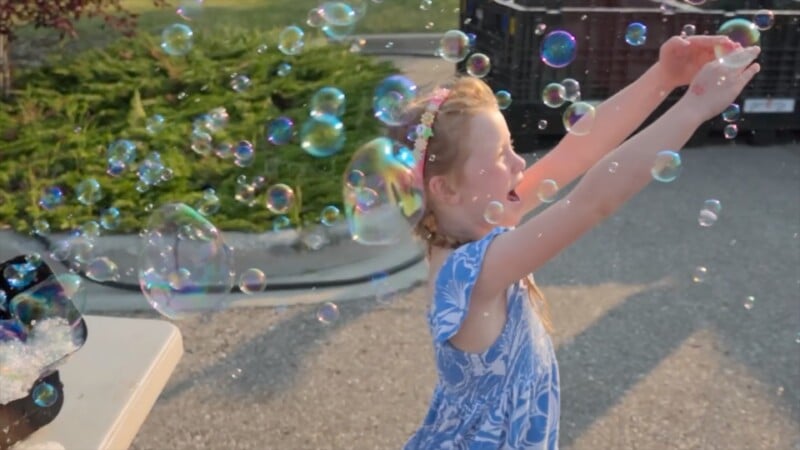 A young girl in a blue floral dress reaches toward a cloud of colorful bubbles floating in the air.