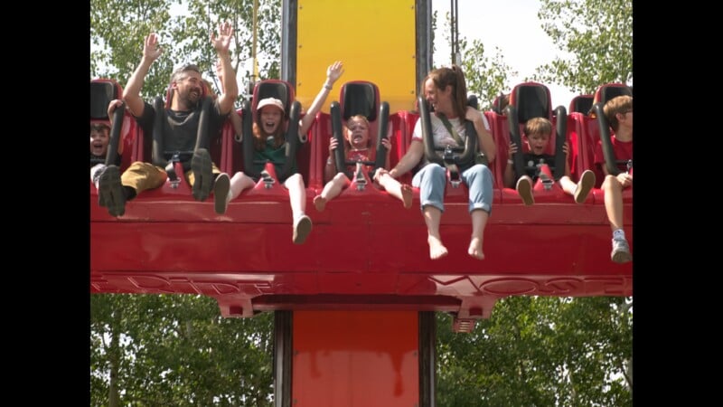 A group of children and adults ride a red drop tower attraction with their arms raised or holding onto the safety restraints.