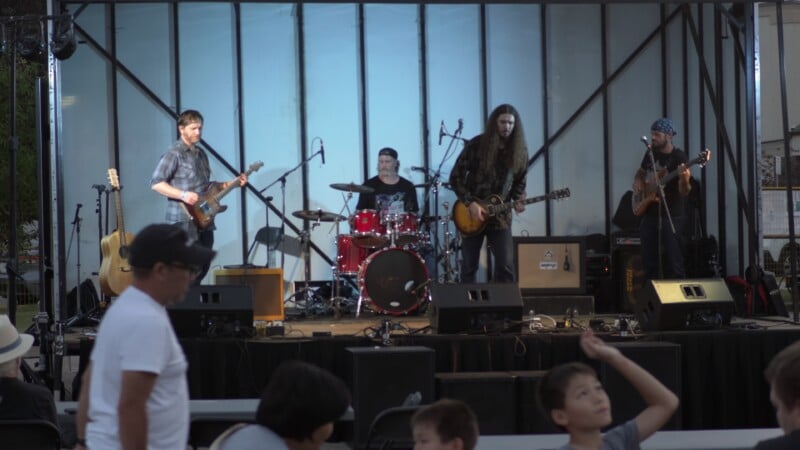 A four-piece band performs on an outdoor stage with a drummer, two guitarists, and a bassist.