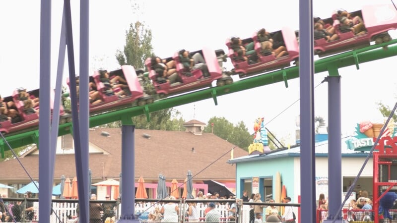 A pink roller coaster train filled with passengers travels along a green track above a park area with food stands and a crowd of people.