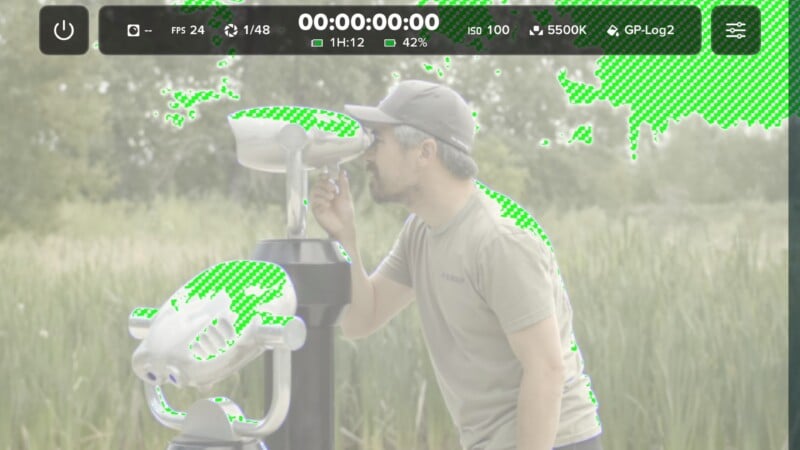 A man looks through a stationary public binocular viewer in a grassy field, with green zebra-stripe exposure warnings overlaid on the bright areas of the frame.