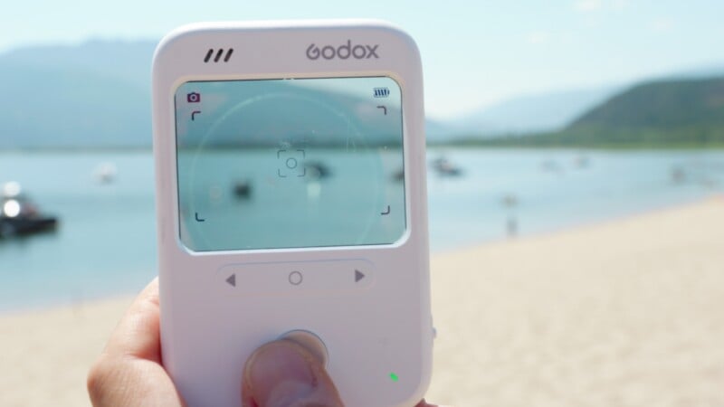 A hand holds a white Godox device with a digital screen displaying a camera viewfinder interface against a blurred beach and water background.