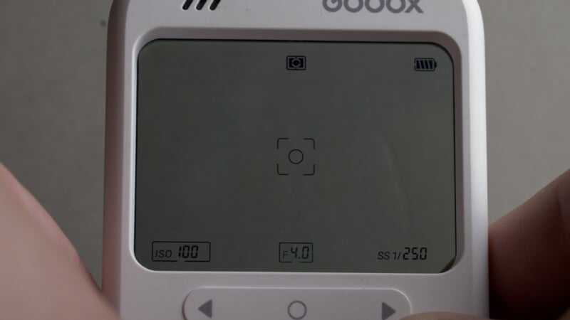 A close-up of a white digital device screen displaying camera settings: ISO 100, F4.0, and SS 1/250, with a central focus bracket icon.