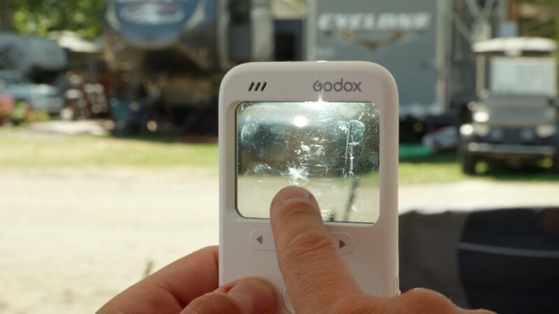 A person holds a white Godox device with a scratched screen in front of an outdoor background featuring a trailer and a golf cart.