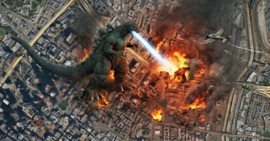 Godzilla stands in the center of a burning city, firing a blue atomic breath beam at the ground while a fighter plane flies nearby.