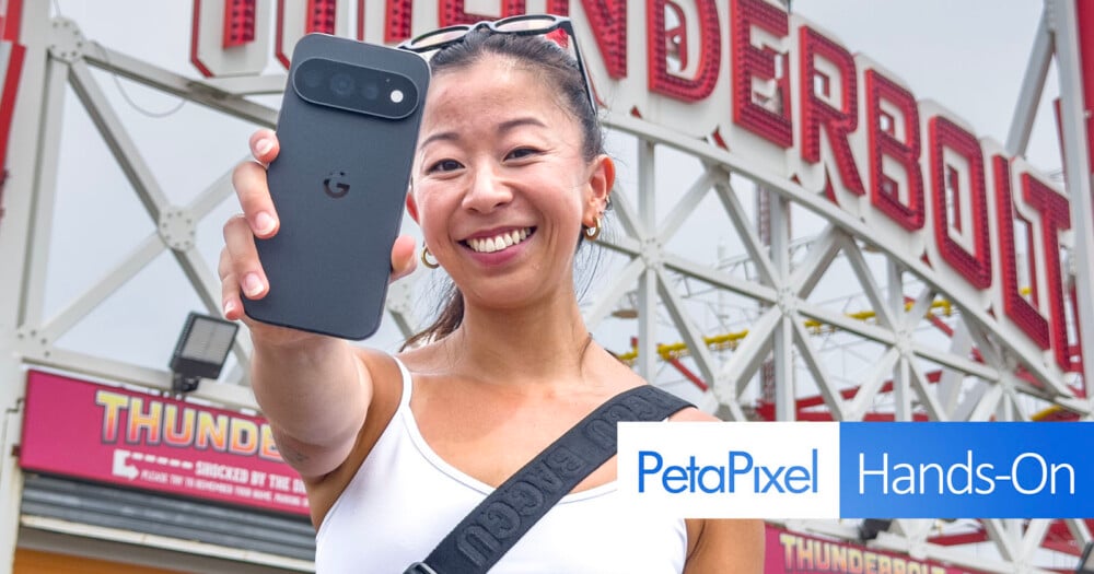 A smiling woman holds a dark smartphone toward the camera in front of a large sign that reads "THUNDERBOLT.