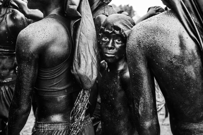 A young child with a dark, painted face and head covering stands between two adults in the rain, all with wet, glistening skin.