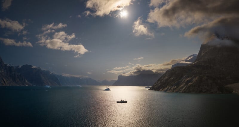 A small boat floats on a calm, sunlit body of water surrounded by towering, rocky mountains and scattered icebergs under a cloudy sky.