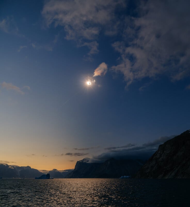 A solar eclipse occurs over a dark, mountainous landscape and a calm sea with icebergs under a twilight sky.