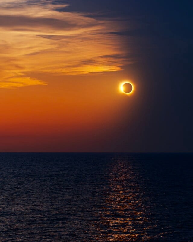 A solar eclipse hangs in a sunset sky above a dark ocean, with the sun's reflection shimmering on the water's surface.