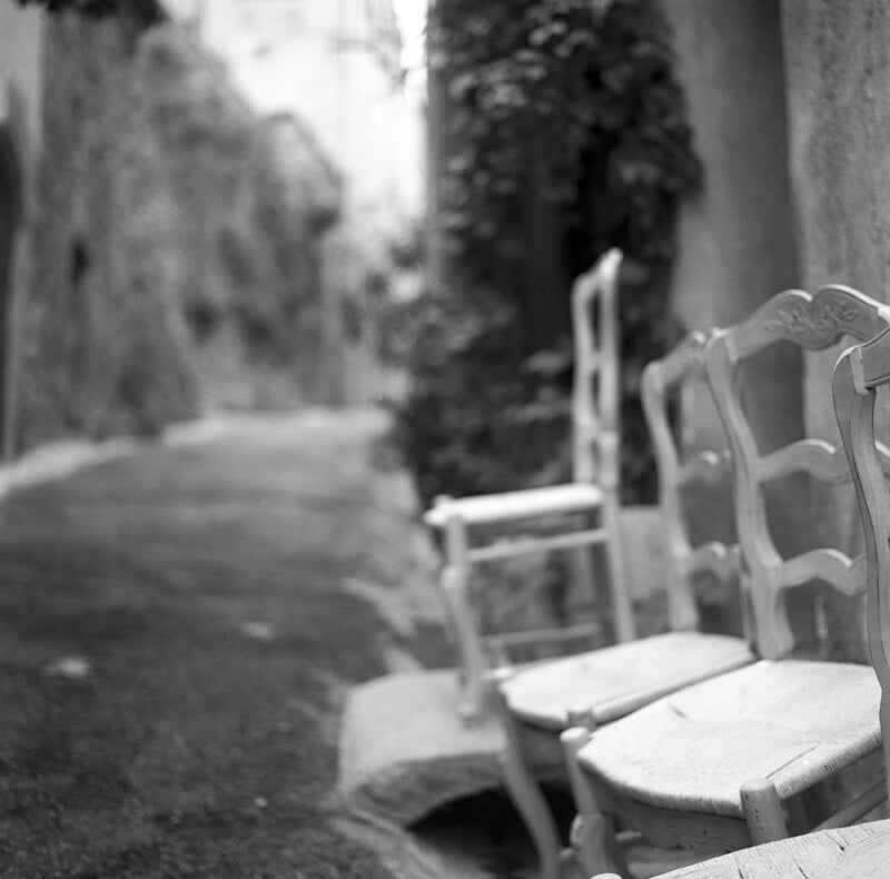 Three wooden chairs with woven seats are lined up along a stone wall in a narrow, blurred alleyway.
