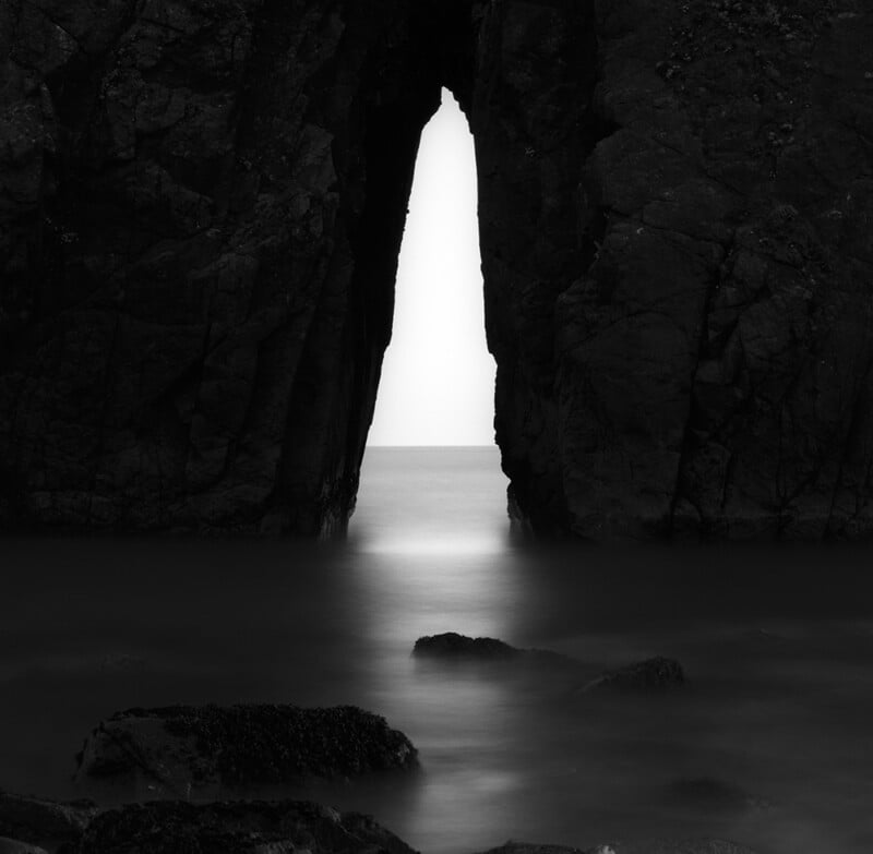 A black and white long-exposure shot of a narrow sea arch framing a bright, hazy horizon over calm water with dark rocks.