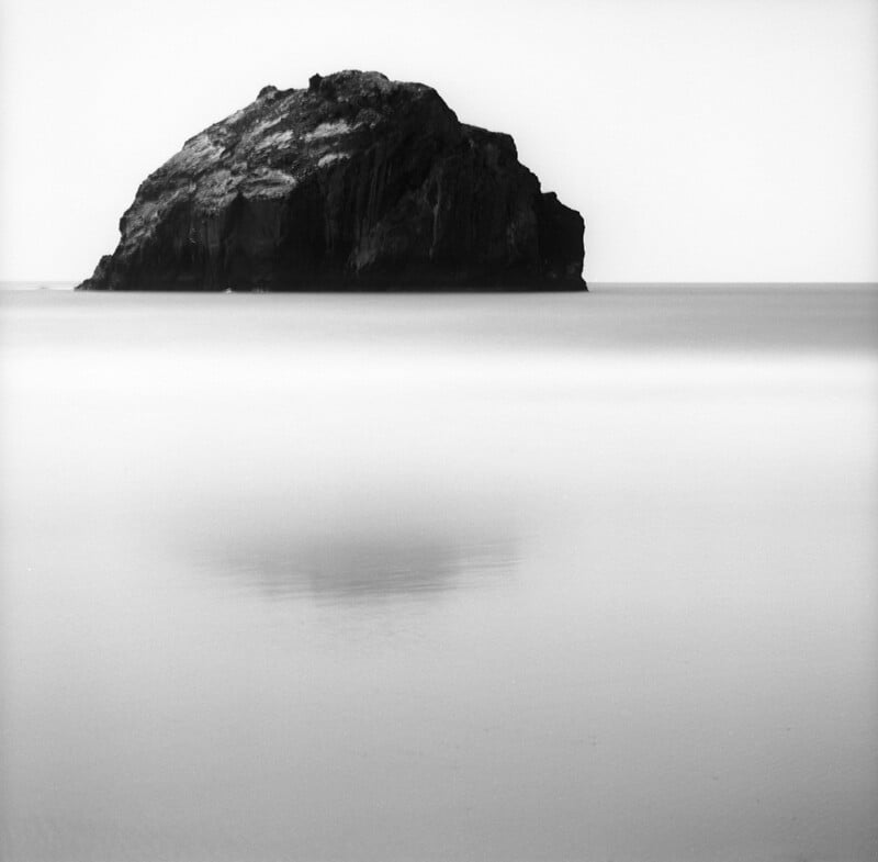 A large, dark rock formation sits in the middle of a calm, misty sea under a bright, featureless sky.